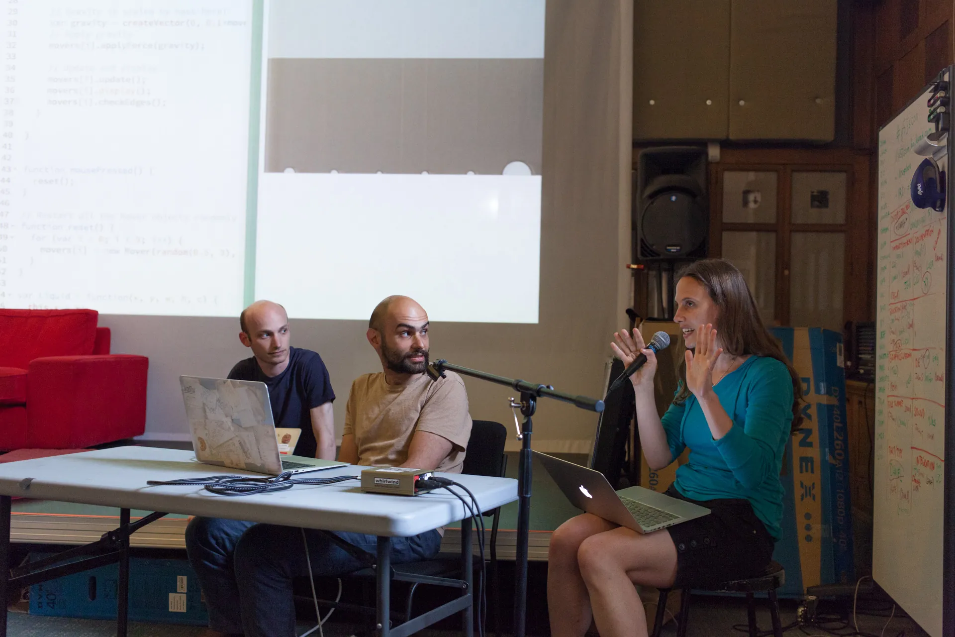 Woman expressively speaks into a microphone while two male collaborators look on