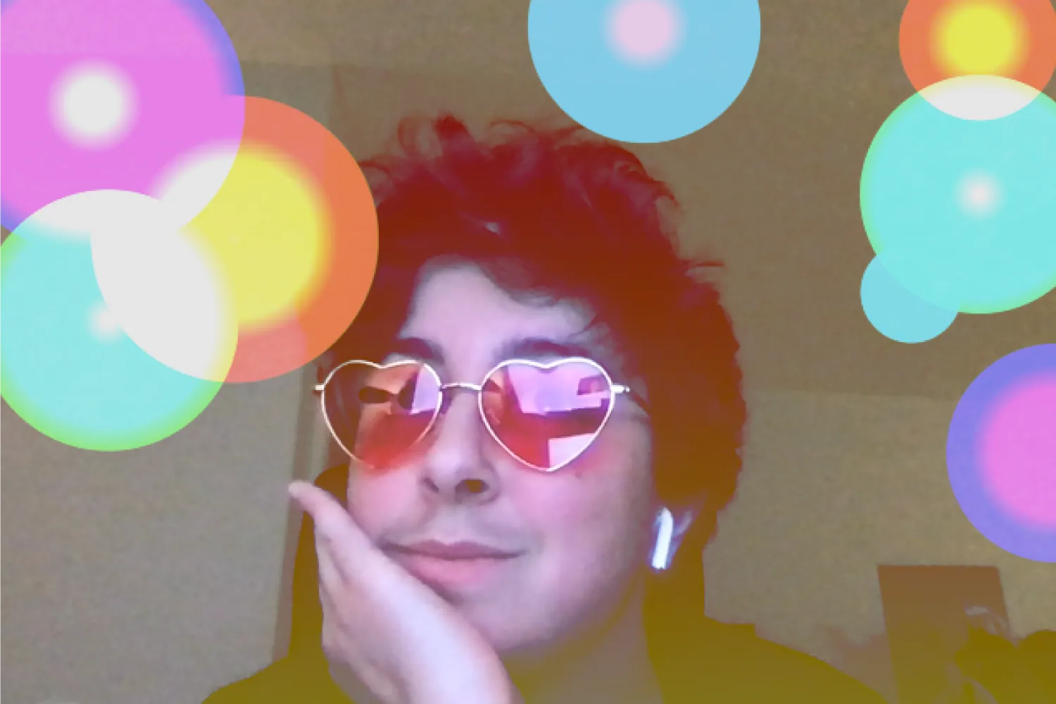 A webcam snapshot of a white person with short dark curly hair wearing pink heart shaped glasses posing for a picture. There is a red to yellow linear gradient filter on top of the photo. Colorful circles are scattered on top of the image with a color gradient overlay. The circles have randomized radial color gradients inside them to mimic a camera's lens flare. Some color gradients are pink to purple, cyan to green, or yellow to orange.