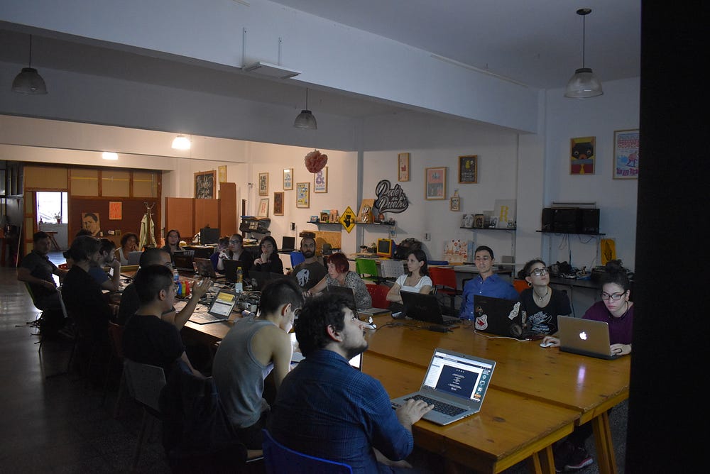 A group of 20 people sitting on a large shared table with their laptops looking at a projected screen.