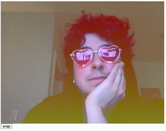 A white person with short dark curly hair wearing pink heart shaped glasses posing with his hand under his chin, with a vertical linear red to yellow gradient applied over the photo as a camera filter. Because of the LIGHTEST blendMode, the colorful gradient is especially visible on his dark hair and dark shirt.