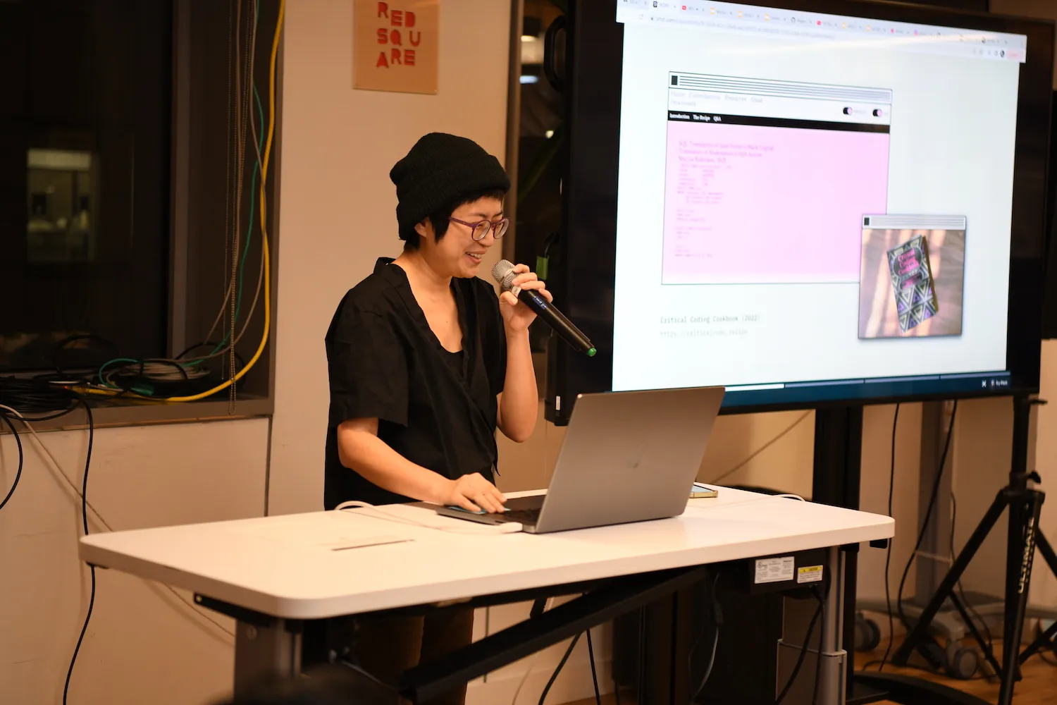 Xin Xin holding a mic next to a big t.v. screen that has an image and text of their work.