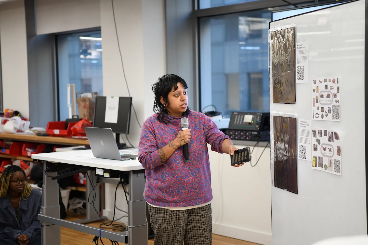 Aarati Akkapedi holding a mic next to a big whiteboard that has a collection of printed work by them.